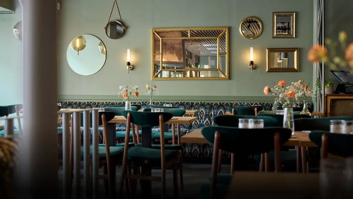 Brown dining tables inside the restaurant. Green chairs around the tables. In the background, a green wall with mirrors and two lamps.